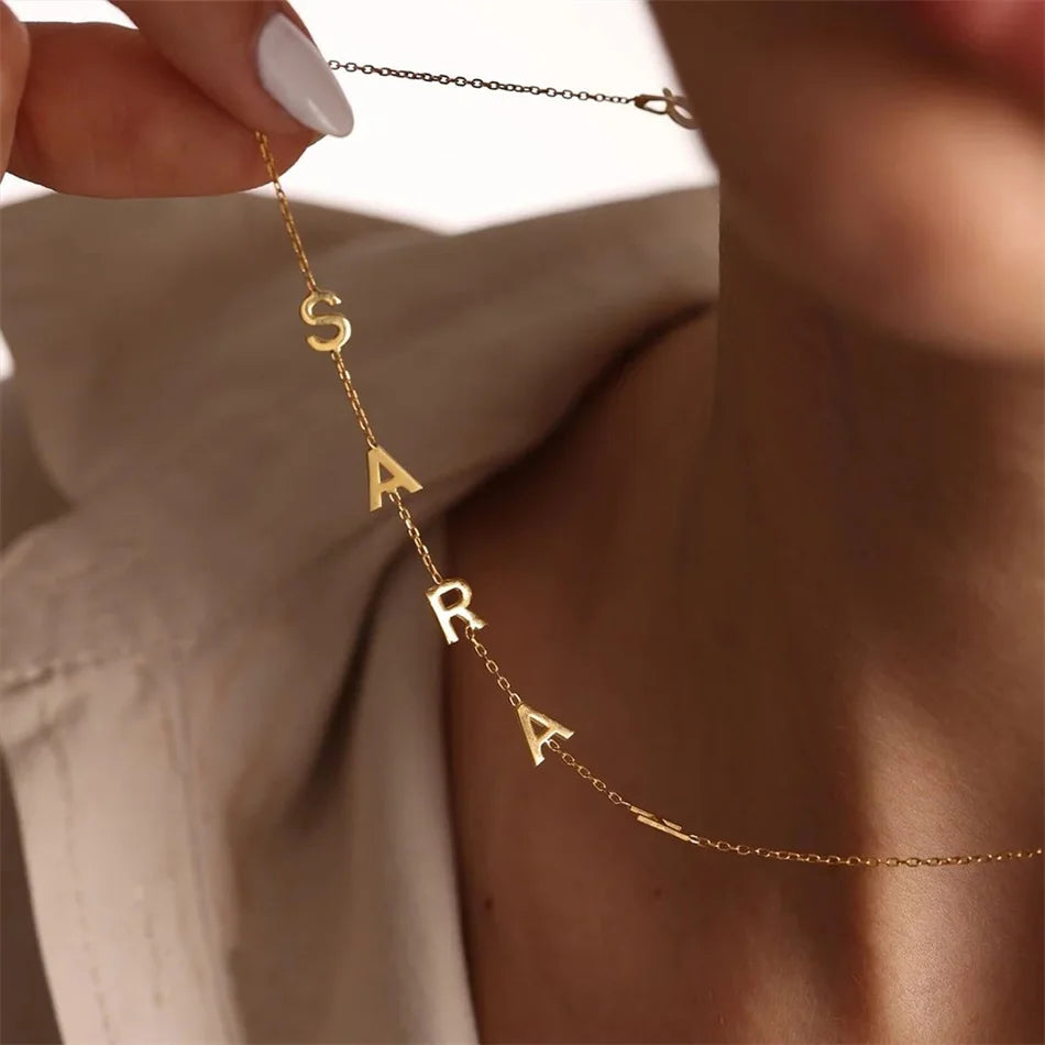 A gold chain necklace spelling out 'SARA' in raised letter charms, held by a hand and worn around a person's neck, shown against a soft beige background.