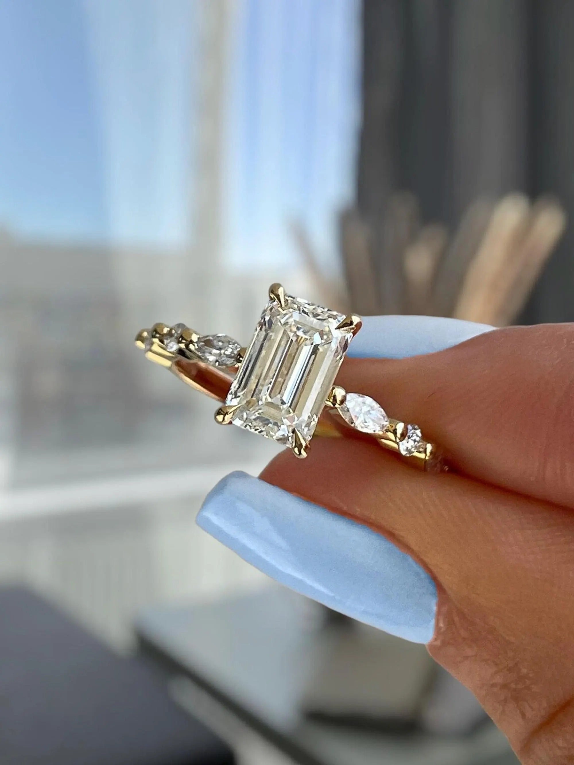 Gold ring with a large diamond held by a hand with light blue nail polish.