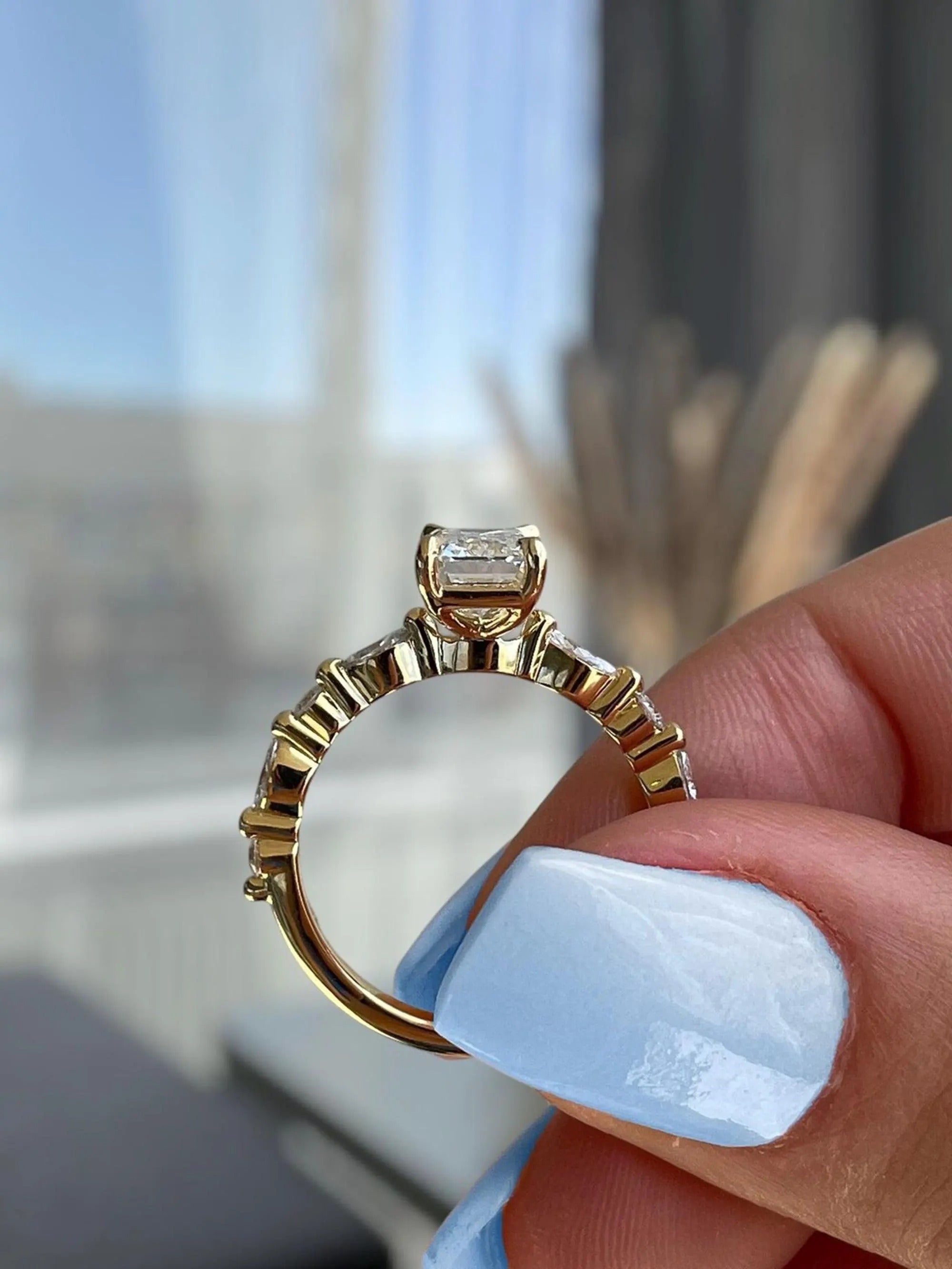 Gold ring with a diamond held by a hand with light blue nail polish, blurred indoor background